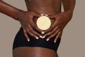 A black woman holding a cocoa butter lotion bar against her mid-section. 